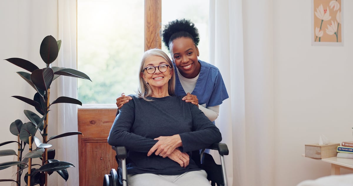 A Black female caregiver smiles while standing behind a white-haired senior woman in a wheelchair at home