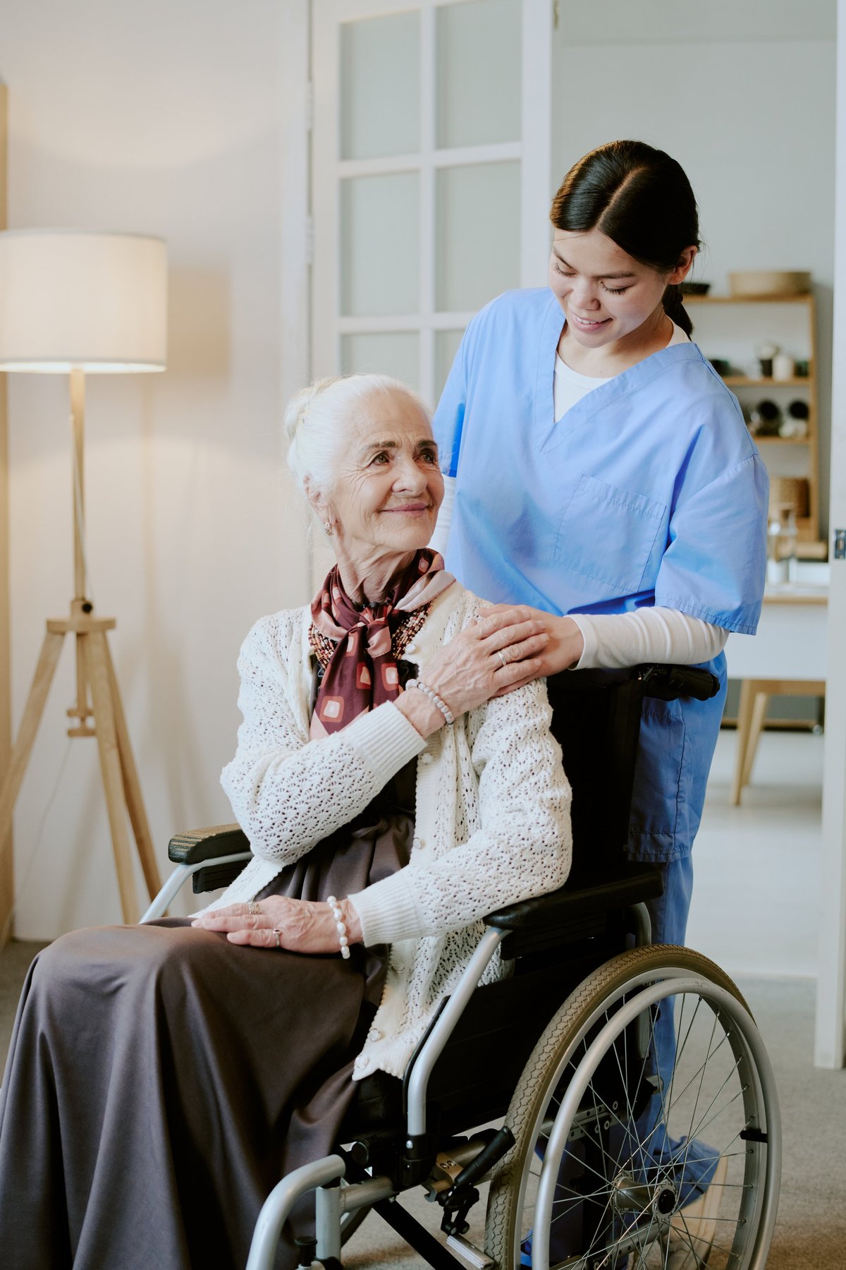A smiling caregiver stands behind a senior woman in a wheelchair