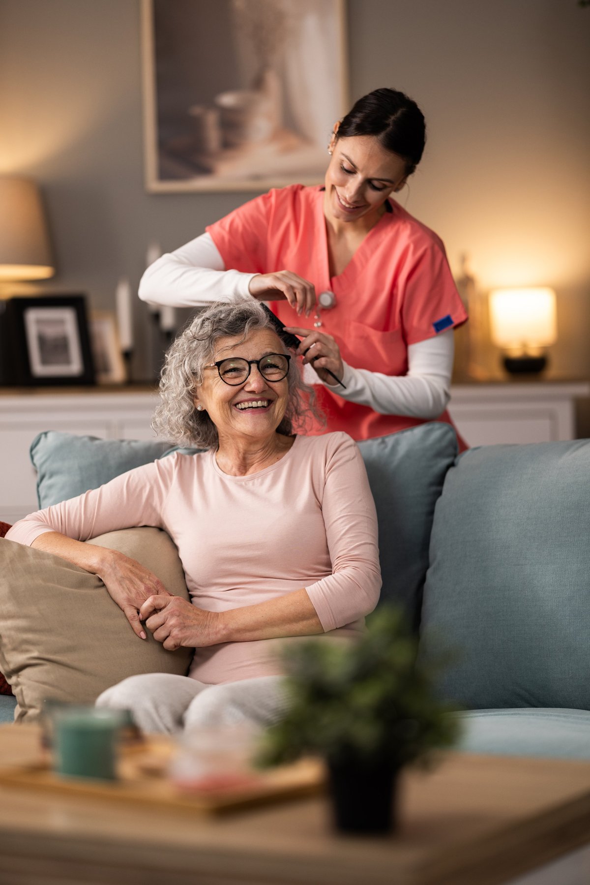 A cheerful caregiver combs a senior woman's hair in the living room
